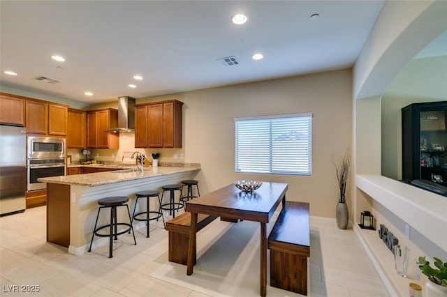 kitchen with light stone counters, visible vents, a peninsula, appliances with stainless steel finishes, and wall chimney exhaust hood