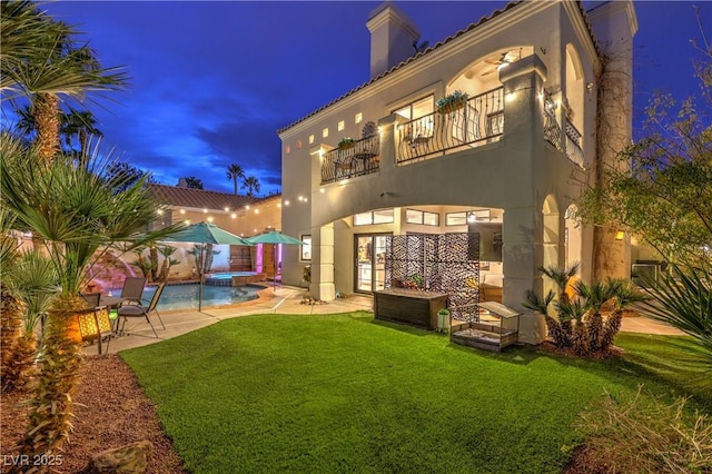 back of property featuring a balcony, a pool with connected hot tub, stucco siding, a patio area, and a lawn