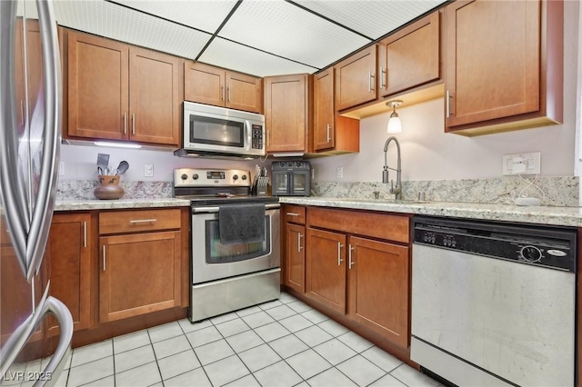 kitchen with light stone counters, light tile patterned floors, brown cabinetry, a sink, and appliances with stainless steel finishes