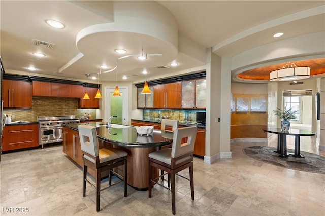 kitchen with range with two ovens, an island with sink, dark countertops, a kitchen breakfast bar, and brown cabinets