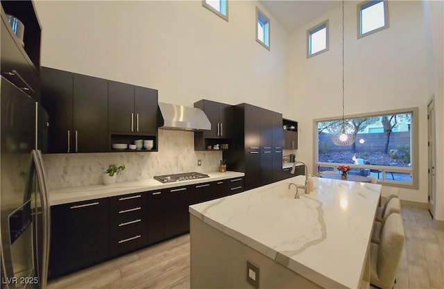 kitchen with wall chimney range hood, pendant lighting, light wood-type flooring, appliances with stainless steel finishes, and dark cabinets