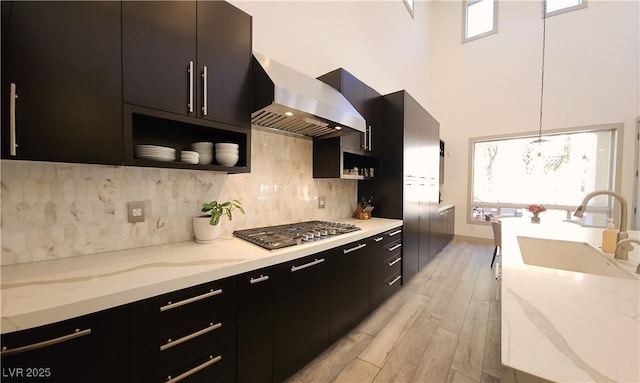 kitchen featuring a sink, tasteful backsplash, wall chimney range hood, dark cabinets, and stainless steel gas cooktop