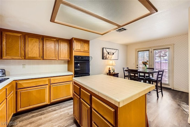 kitchen featuring brown cabinets, black oven, tile countertops, light wood-style floors, and wallpapered walls