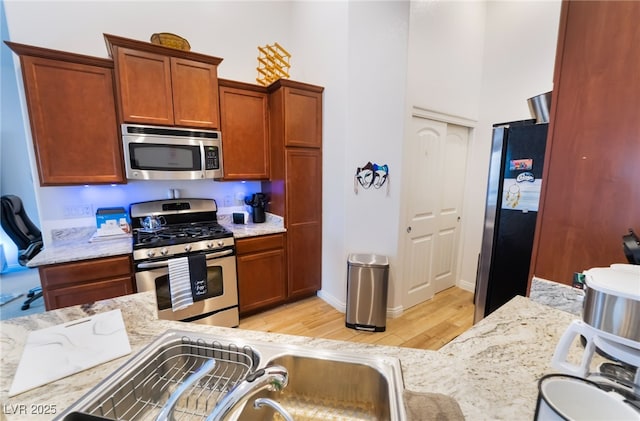 kitchen with brown cabinetry, light wood finished floors, a sink, a towering ceiling, and appliances with stainless steel finishes