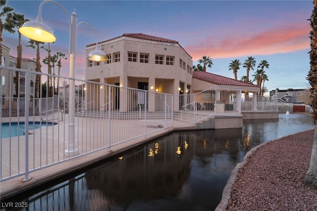 back of house at dusk with a fenced in pool, fence, a water view, a tiled roof, and stucco siding