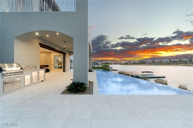 view of swimming pool featuring grilling area, a water view, exterior kitchen, a patio area, and a ceiling fan