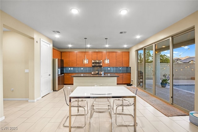 kitchen featuring visible vents, an island with sink, dark countertops, stainless steel appliances, and brown cabinetry