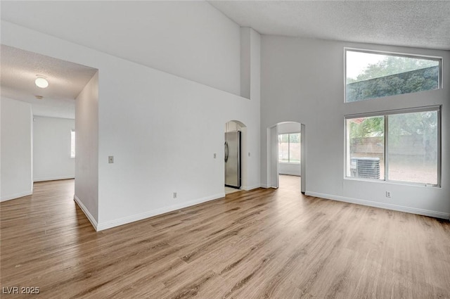 unfurnished living room with light wood-type flooring, a textured ceiling, arched walkways, baseboards, and a towering ceiling