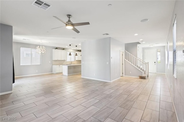 unfurnished living room with stairway, visible vents, baseboards, a sink, and ceiling fan with notable chandelier