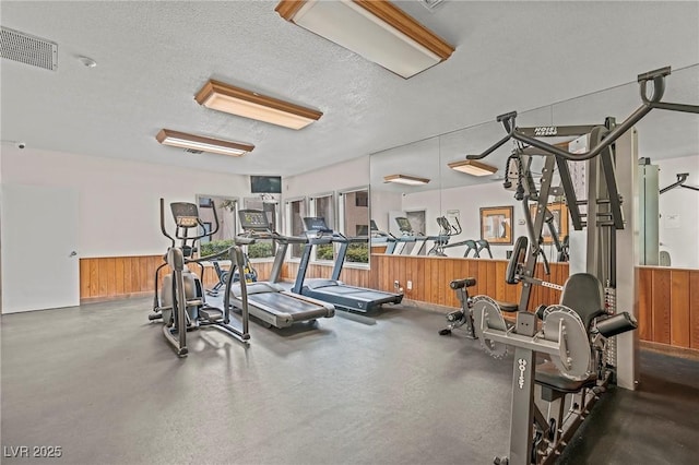 workout area with visible vents, wooden walls, a textured ceiling, and wainscoting