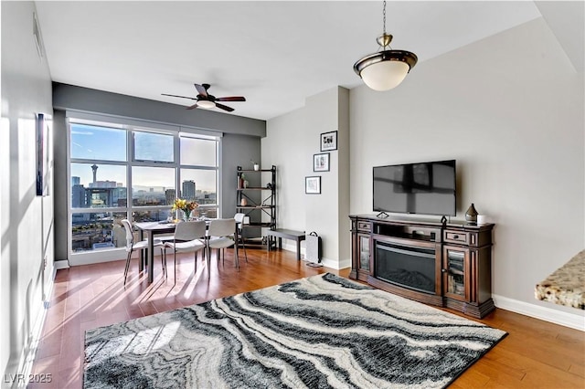 living area with a glass covered fireplace, wood finished floors, baseboards, and ceiling fan