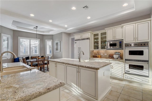 kitchen featuring visible vents, an island with sink, a sink, built in appliances, and a raised ceiling