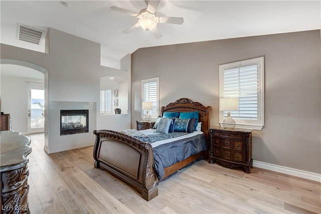 bedroom featuring visible vents, a multi sided fireplace, light wood-type flooring, vaulted ceiling, and arched walkways