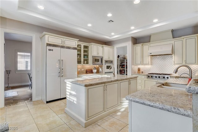 kitchen featuring built in appliances, light stone counters, range hood, a raised ceiling, and a sink