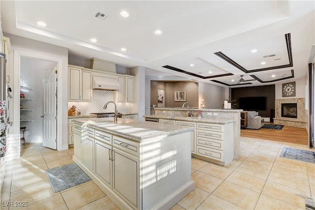 kitchen featuring a fireplace with raised hearth, an island with sink, light stone counters, light tile patterned flooring, and cream cabinetry