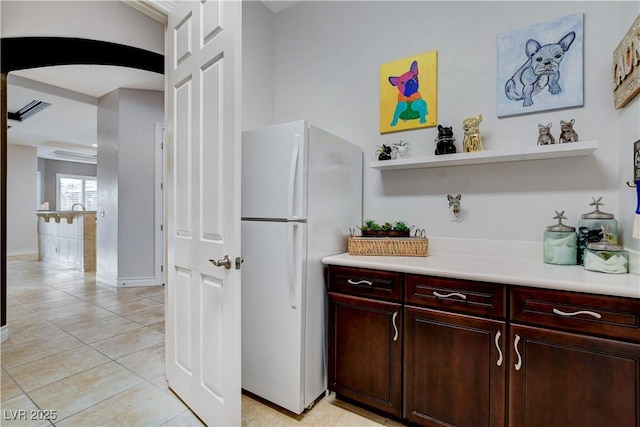 kitchen featuring light tile patterned floors, arched walkways, freestanding refrigerator, light countertops, and dark brown cabinets