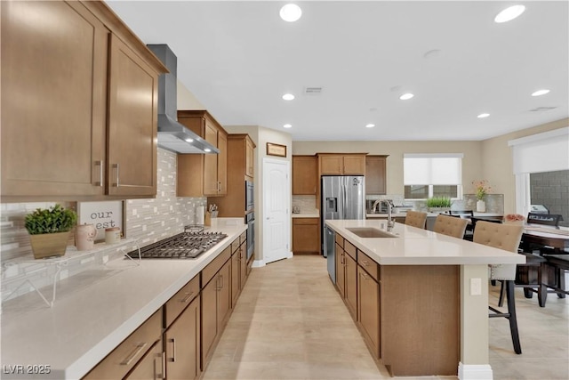 kitchen with a breakfast bar, a center island with sink, a sink, appliances with stainless steel finishes, and wall chimney range hood