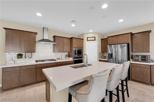 kitchen featuring a center island with sink, a breakfast bar area, appliances with stainless steel finishes, wall chimney exhaust hood, and a sink