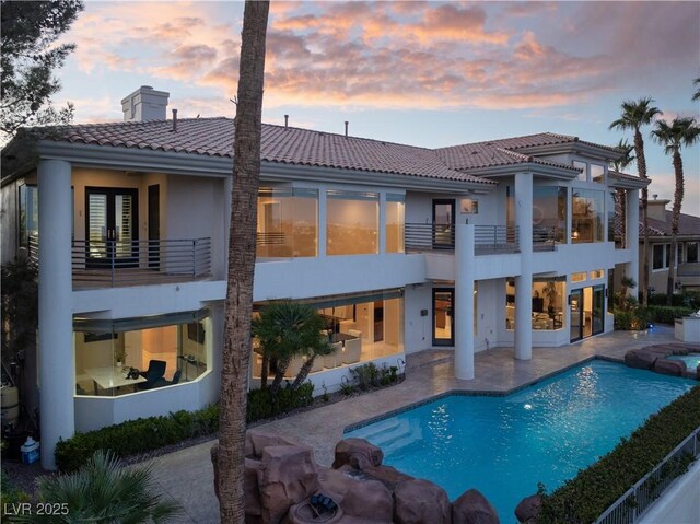 back of house at dusk featuring a tile roof, a patio, french doors, a balcony, and a chimney