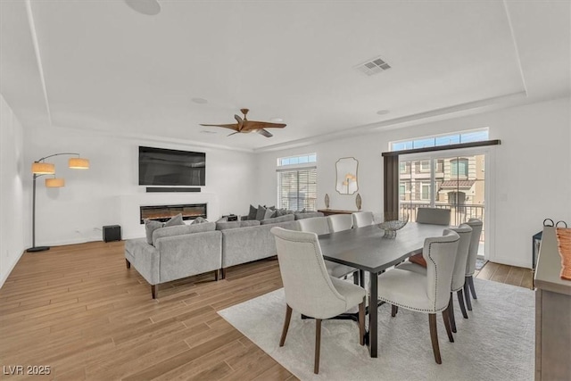 dining room with visible vents, ceiling fan, light wood-type flooring, a glass covered fireplace, and a raised ceiling