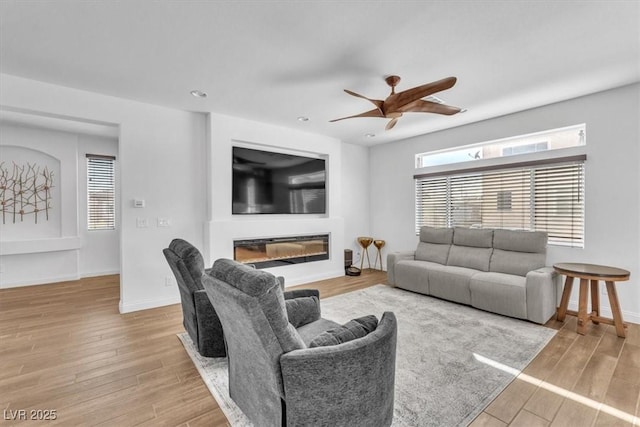 living area with light wood finished floors, a glass covered fireplace, ceiling fan, and baseboards