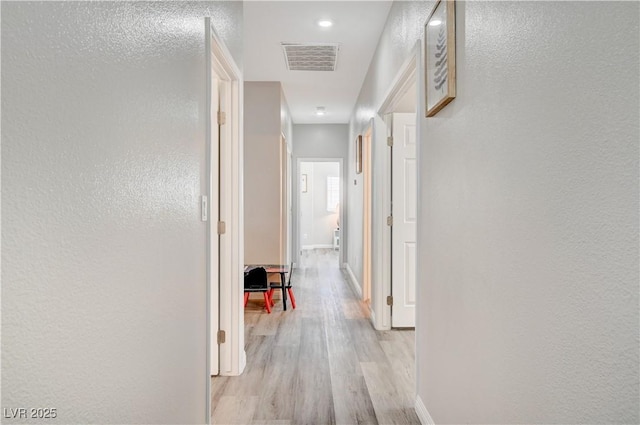 hallway with visible vents, baseboards, light wood-style floors, and a textured wall