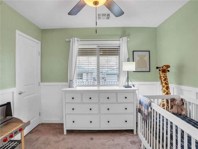 bedroom with a ceiling fan, visible vents, wainscoting, a crib, and light carpet