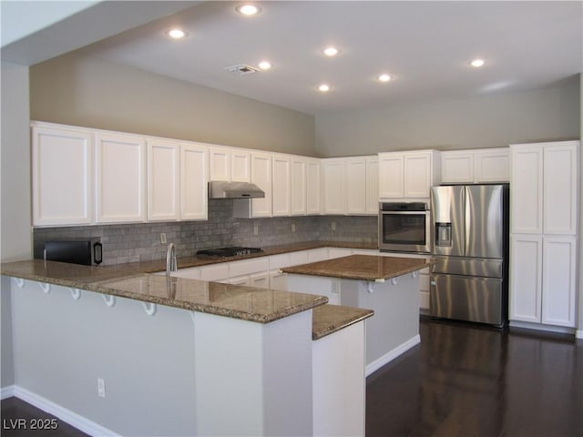 kitchen featuring dark stone countertops, a peninsula, stainless steel appliances, white cabinets, and under cabinet range hood