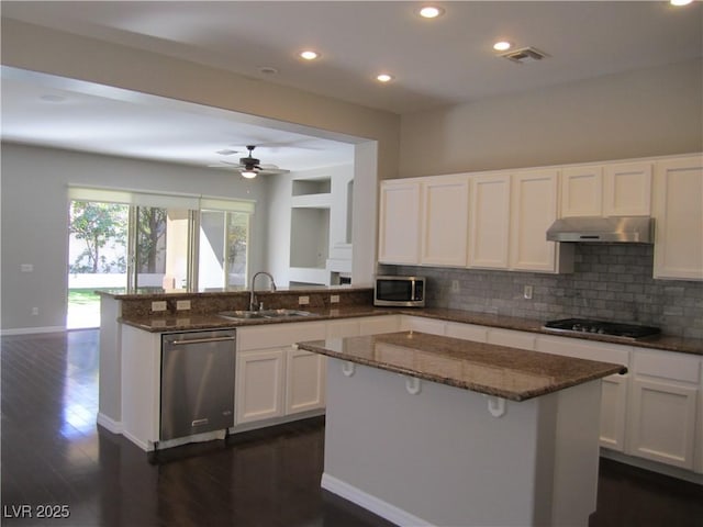 kitchen featuring under cabinet range hood, dark stone countertops, a sink, appliances with stainless steel finishes, and a peninsula