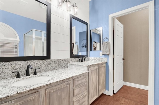 bathroom featuring wood finished floors, baseboards, double vanity, a stall shower, and a sink