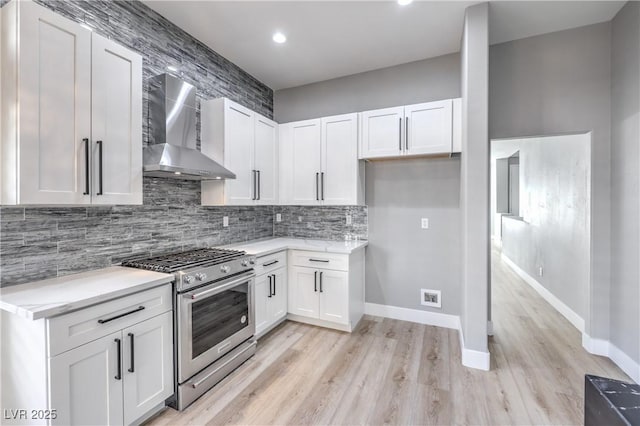 kitchen with wall chimney range hood, backsplash, white cabinetry, stainless steel stove, and light wood finished floors