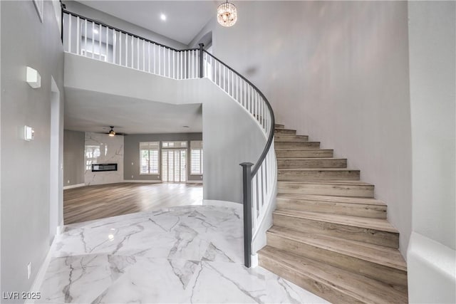 stairs with ceiling fan with notable chandelier, wood finished floors, a high ceiling, a premium fireplace, and baseboards