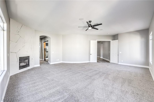 unfurnished living room featuring a ceiling fan, arched walkways, carpet floors, a premium fireplace, and baseboards