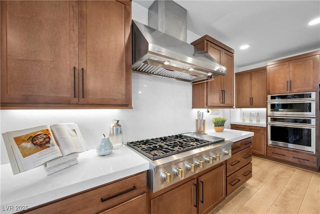 kitchen with brown cabinetry, wall chimney exhaust hood, light wood-style floors, and appliances with stainless steel finishes