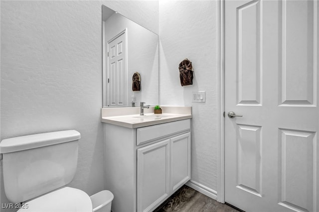 half bathroom with toilet, wood finished floors, baseboards, vanity, and a textured wall