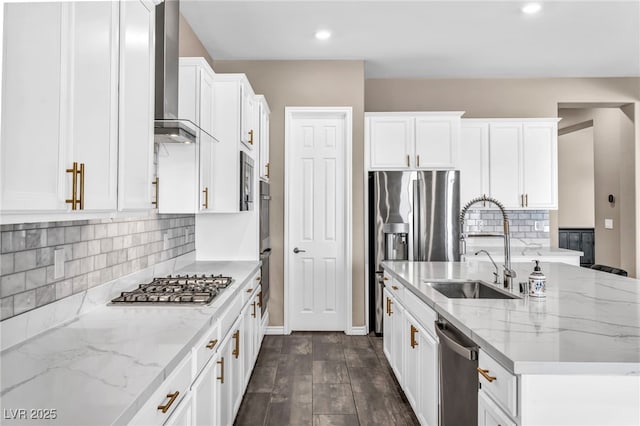 kitchen with a center island with sink, a sink, white cabinetry, stainless steel appliances, and wall chimney range hood