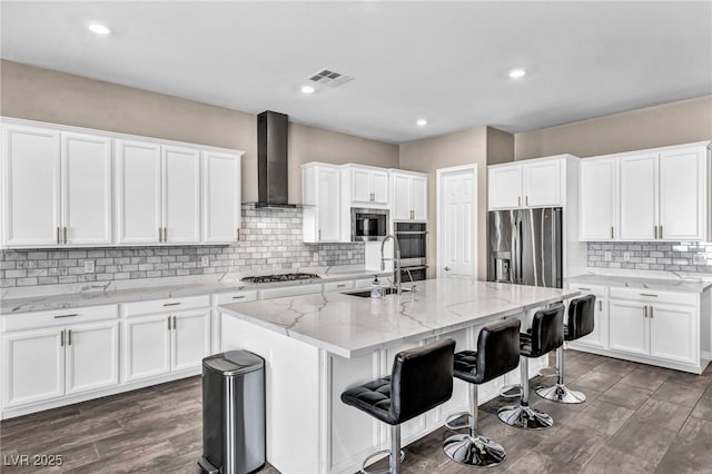 kitchen featuring visible vents, a center island with sink, a sink, stainless steel appliances, and wall chimney exhaust hood