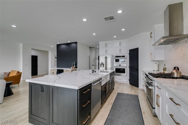 kitchen featuring visible vents, a large island with sink, white cabinets, stainless steel appliances, and wall chimney exhaust hood