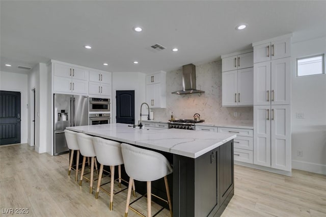 kitchen with visible vents, a kitchen island with sink, a sink, stainless steel appliances, and wall chimney range hood