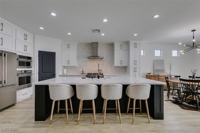 kitchen featuring visible vents, a notable chandelier, stainless steel appliances, wall chimney exhaust hood, and a large island with sink