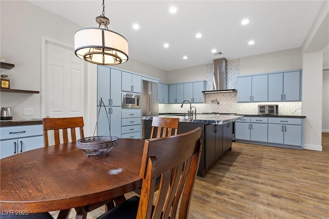 kitchen with a kitchen island with sink, backsplash, wood finished floors, stainless steel appliances, and wall chimney exhaust hood