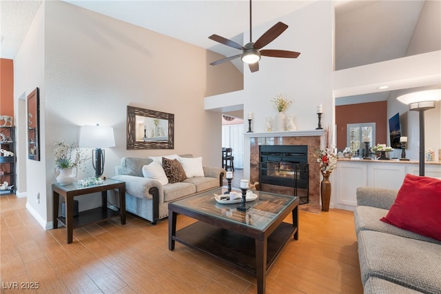 living room featuring light wood-style flooring, a ceiling fan, a tiled fireplace, baseboards, and a towering ceiling