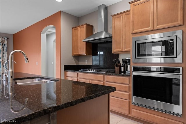 kitchen featuring dark stone counters, light tile patterned floors, stainless steel appliances, wall chimney exhaust hood, and a sink