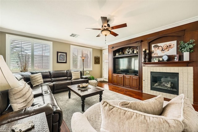 living area featuring wood finished floors, visible vents, ceiling fan, a tiled fireplace, and crown molding