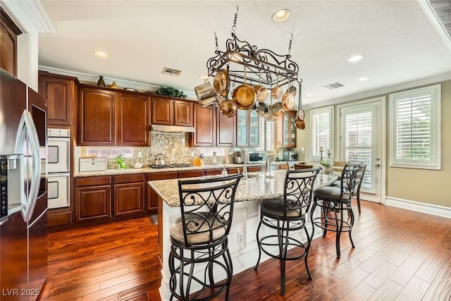 kitchen with dark wood-type flooring, under cabinet range hood, a breakfast bar area, ornamental molding, and appliances with stainless steel finishes