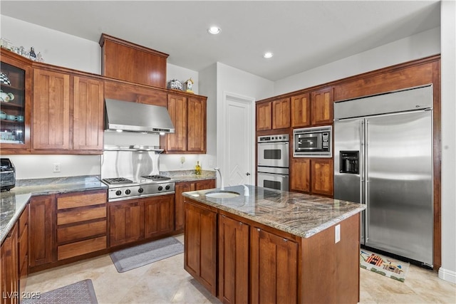 kitchen with a center island, glass insert cabinets, wall chimney range hood, built in appliances, and dark stone counters