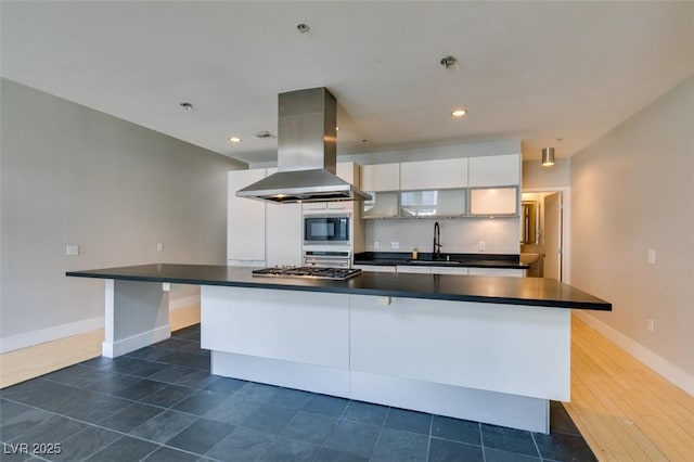 kitchen featuring island exhaust hood, a sink, appliances with stainless steel finishes, white cabinetry, and dark countertops