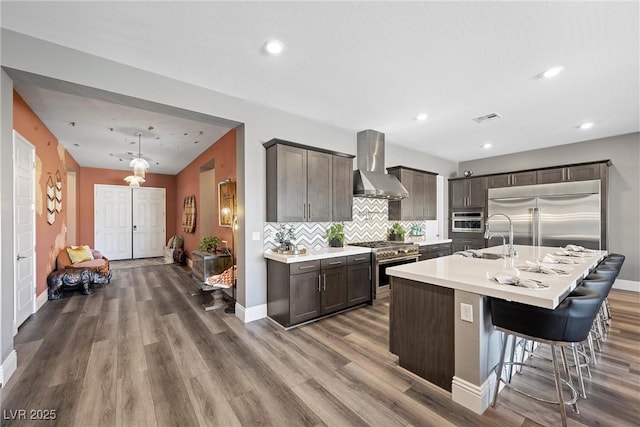 kitchen featuring wall chimney range hood, dark brown cabinetry, decorative backsplash, high end appliances, and a sink