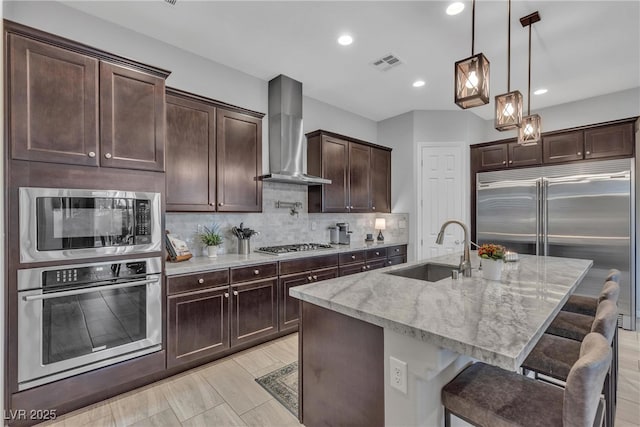 kitchen featuring light stone countertops, a sink, built in appliances, wall chimney exhaust hood, and tasteful backsplash