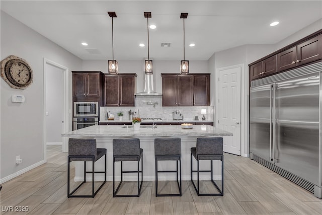 kitchen with visible vents, light stone counters, tasteful backsplash, wall chimney range hood, and built in appliances